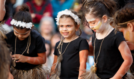 Young Indigenous girls with face paint and wearing black tops and grass skirts stand next to each other. In the background are more people.