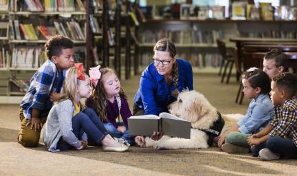 And adult woman sits on the floor holding a book so that children either side of her can see it. In the background are bookshelves.