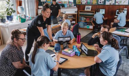 A group of students sit around a table as they work with laptops. A middle-aged woman dressed in black leans on the table while another sits and looks on. In the background are more students, a teacher and classroom paraphernalia.