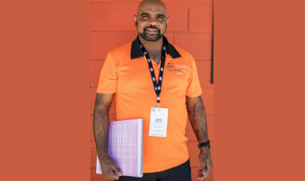 Man standing against brick wall wearing orange 2021 Census shirt and black lanyard holding paperwork.