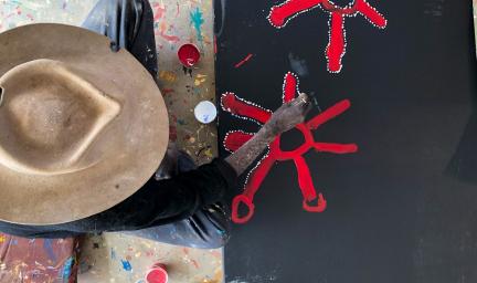 A man in a brown hat and dark shirt sits next to a black canvas on the floor. He is painting red starfish like figures.