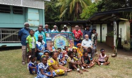 Group of people, some sitting and others standing on grass in front of a building. They are dressed in colourful clothing. In the background are trees.