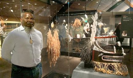 Indigenous man in pale shirt and jeans with a beard, stands next to a yellow mask of draping filaments suspended on wires from above. In the background and panes of glass and more Indigenous masks and items.