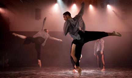 Four male and female dancers on a floor in softly lit room. Two dancers are in action while two others stand and crouch.