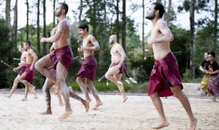 A group of males dressed in maroon loin cloths and holding sticks dance on sand. Behind them are two female dancers in shirts and wraps. In the background are trees.