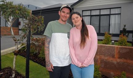 A man wearing a green and white shirt and black shorts is holding a woman wearing a pink long-sleeved shirt and light blue jeans outside a one storey home a tree to their right and a brown fence behind them.