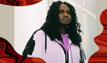 Baker Boy pictured with long black hair wearing a dark pink jumper and a light pink jacket with black outlining with the writing WINNER at the top of the image and the NIMA logo at the bottom left next to the title Artist of the Year Baker Boy.