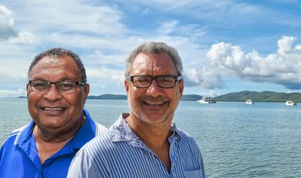 Head and shoulder photo of two men, both in blue shirts and wearing glasses. In the background is a large expanse of water, boats and hills.