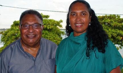 A man in a grey shirt and a woman in a green dress face the camera. In the background are trees and sky.