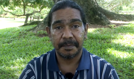 Man in dark blue and white striped shirt sits in grassed area in front of large tree.