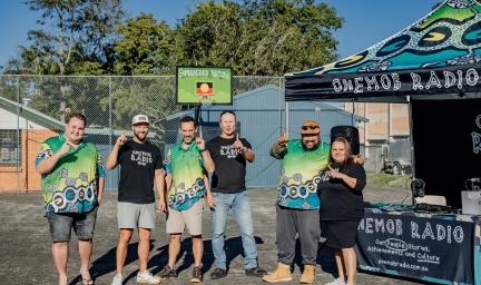 Six people in casual wear stand shoulder to shoulder on a hard surface outdoors. Next to them is a table with a banner displaying the words OneMob Radio. In the background is a building.