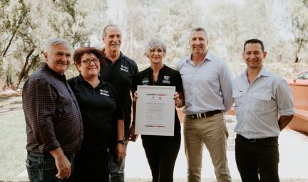 Six people wearing casual clothing stand shoulder to shoulder. In the middle a woman holds up a framed certificate.