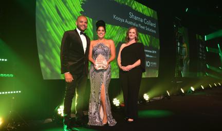 Three people dressed in formal wear and one of them holding an award, stand on a stage and in front of a projected image. In the background are also footlights.