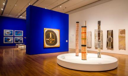 A gallery with a beige ceiling and shiny board floor features two blue walls. In the gallery are different pieces of Indigenous art.