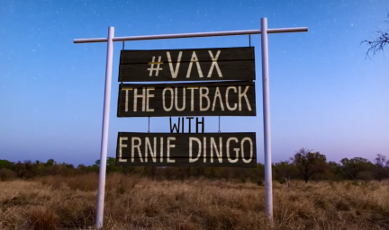 A sign reads # Vax the outback with Ernie Dingo. In the background there is a sunset with dry grass and scrub below the sign in the foreground.