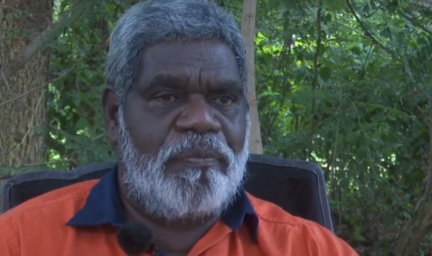 Man in orange shirt with dark blue collar, sits in black chair in front of trees.