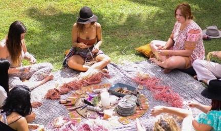 A group of women sit on grass and around a blanket upon which is thread and other items necessary for weaving.