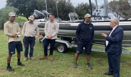 Five men stand in front of a large boat on a trailer on grass. In the background are trees. Four men are dressed in ranger uniforms and the fifth is wearing a suit.