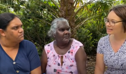 Three women sit next to one another in front of several trees and bushes in the background.