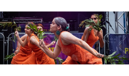 Three women dressed in orange costumes and wearing white paint on arms and faces, move around a stage holding leafy branches.