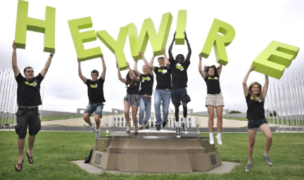 A group of youth and young adults hold large yellow letters above their heads as some of them leap from a seat. The letters spell the word Heywire. In the background is a building and flagpoles.