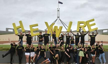 Two rows of youth in casual dress and wearing black t-shirts stand atop and in front of a wall. Some of those on the wall hold large yellow letters which spell: heywire.