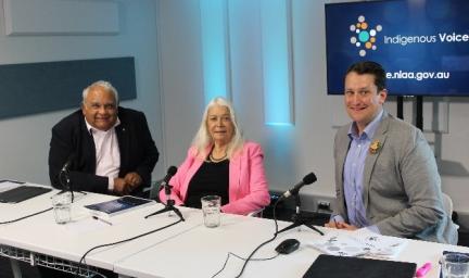 Two men and one woman sit at a table, each with a microphone in front of them. On the table are books, papers and other materials.