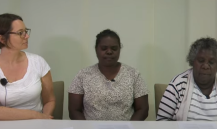 Three woman sit at a white desk, in front of a grey wall. The woman on the left is wearing a white t-shirt. The woman in the middle is wearing a brown and white patterned t-shirt. The woman on the right is wearing a grey and white striped top.