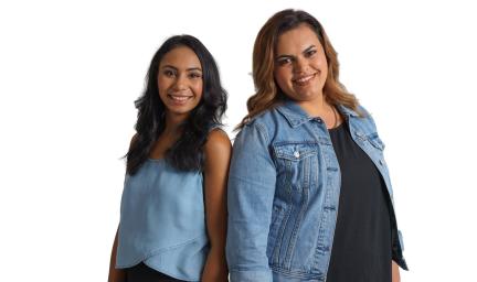 Two young women stand side by side smiling. One woman is wearing a blue top, black pants and has long black hair. The other woman is wearing a blue denim jacket, black top and has brown hair.