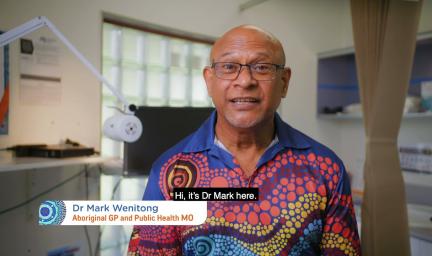 A man sitting in doctor’s surgery wearing a brightly coloured polo shirt and glasses. Words read: Doctor Mark Wenitong Aboriginal GP and Public Health MO. Hi it’s Dr Mark.