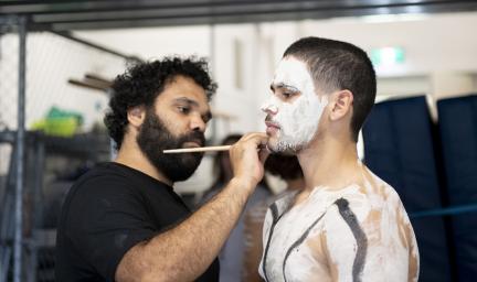 A man in black t-shirt is painting the upper body of another man. He has paint on his face, shoulders and chest.