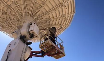 A man in workwear stands on a secure platform while he works below a large white satellite dish.