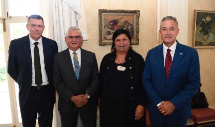 Three men in dark suits and one woman in dark dress and jacket stand shoulder to shoulder in a room. Behind them are paintings, a curtain, a doorway and chairs.