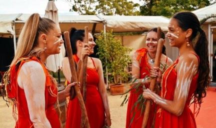 Four women in red dressed and with traditional designs painted on the face and arms talk with each other. In the background are some shelters and potted trees.