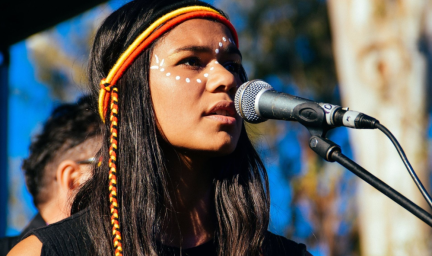 A young Indigenous woman with white dots painted across her face and wearing a black, red and yellow band around her head and long black hair stands in front of a microphone. In the background are trees and blue sky.