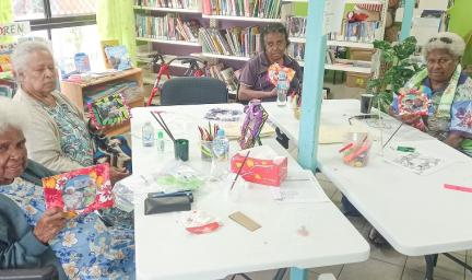 Four elderly women sit around two tables and face the camera. On the table are paint brushes, tissues and more items. In the background are books, boxes and furniture.